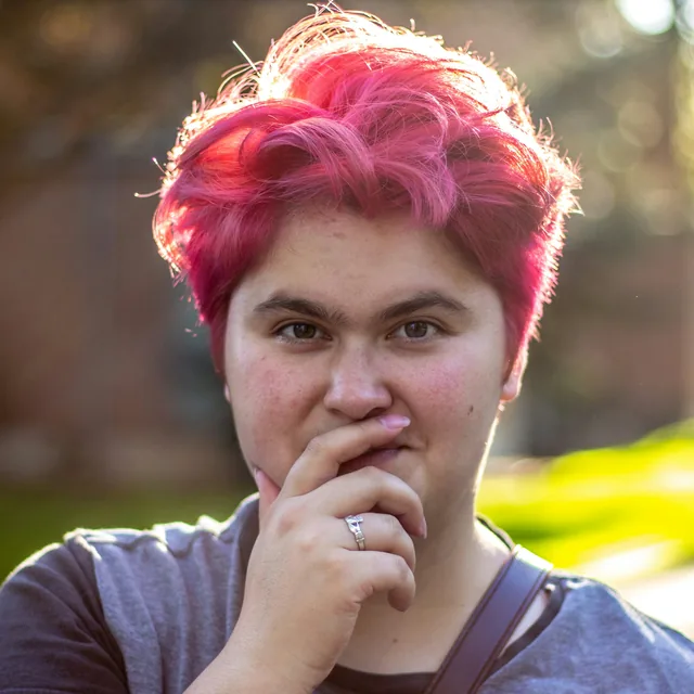 A young adult with bright pink hair, outdoors in evening light
